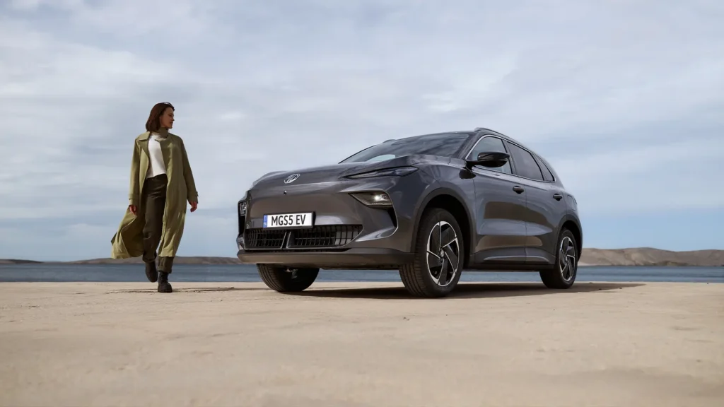 Kvinne i frakk går mot grå bil på sand, skyer og åser.