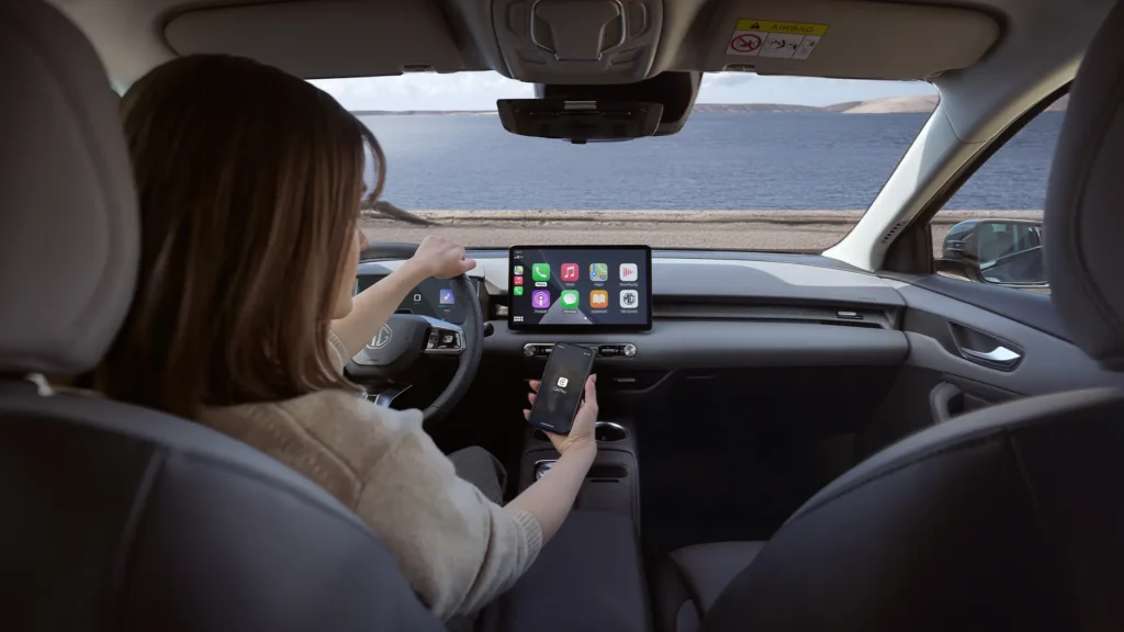Person i bilfører sete ved vann interagerer med skjerm, holder mobil.
