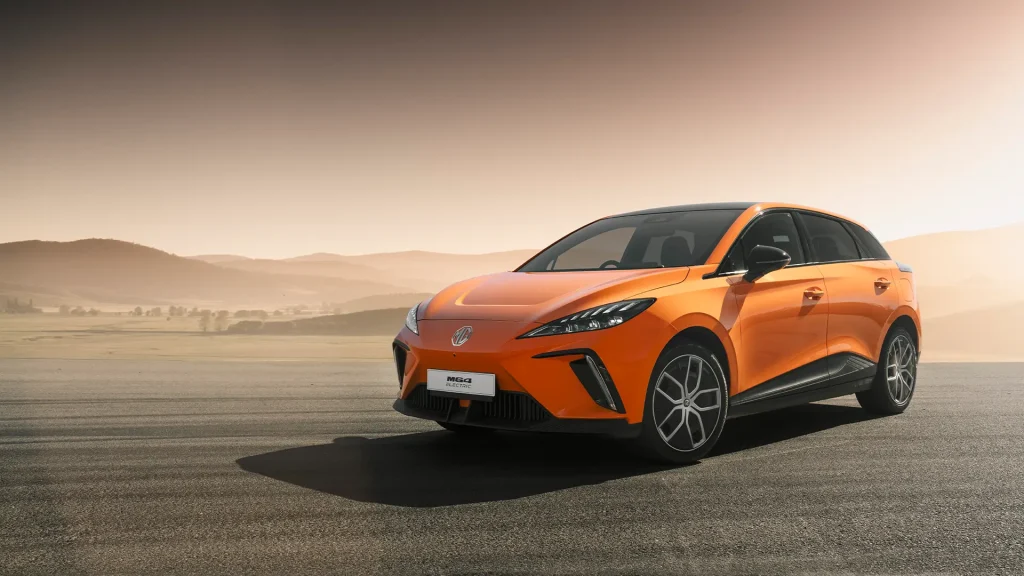 Oransje elbil parkert på asfalt, med åser og disig himmel bak.