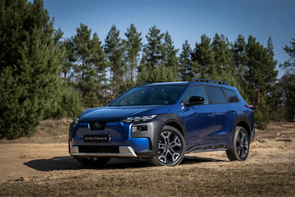 Blå Subaru E-Outback parkert på grusvei med trær og klar himmel.