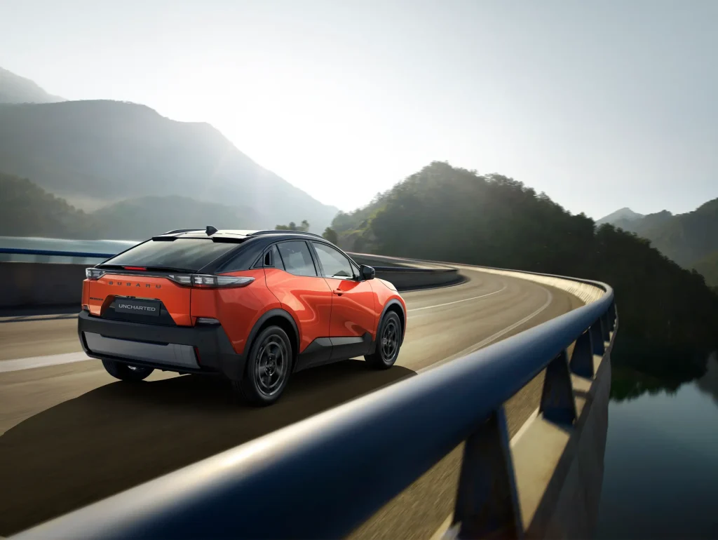 Oransje SUV kjører på svingete fjellvei ved vann og bakker.