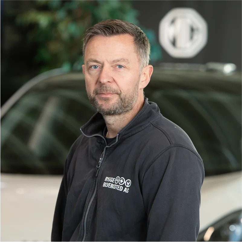 Mann med skjegg står foran hvit bil innendørs.