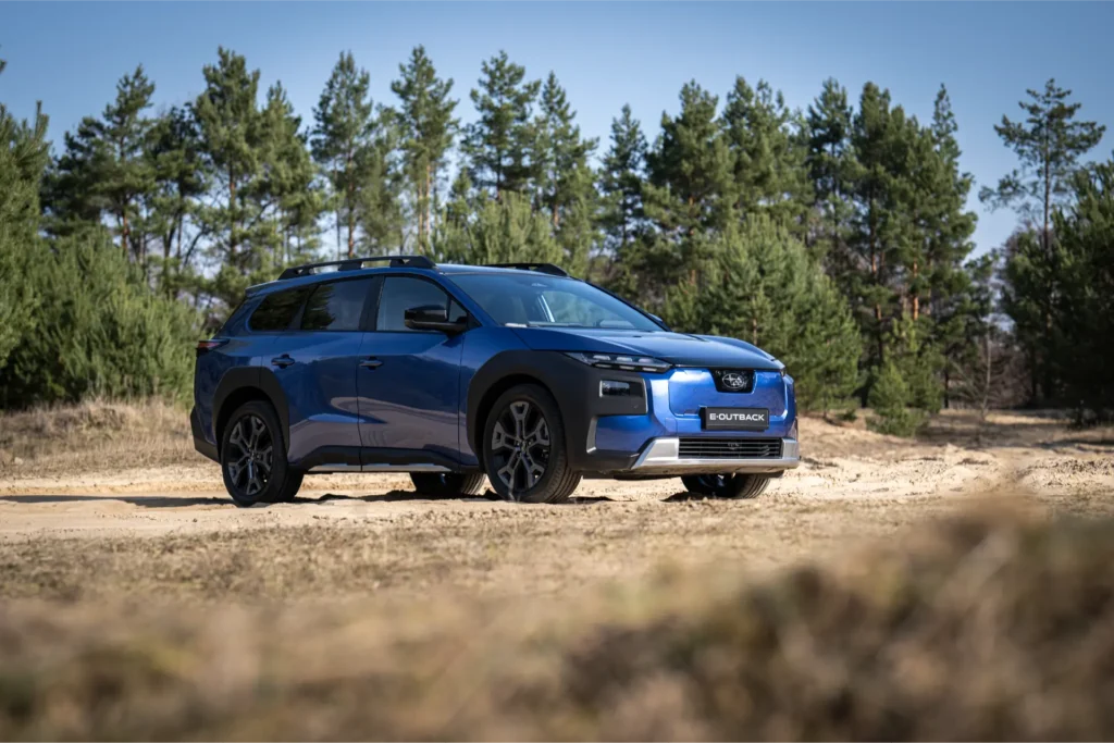 En blå Subaru E-Outback står parkert på en grusvei foran grønne trær.